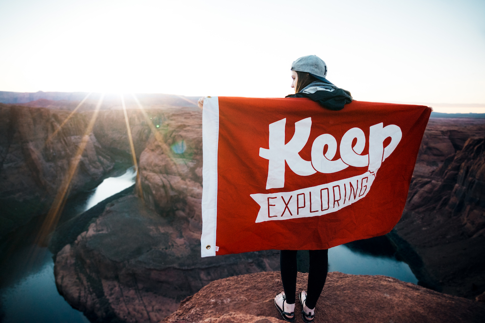 image of a man on a mountain top with a flag that reads "Keep Exploring"